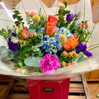 Bright Delight - A gloriously colourful handtied bouquet in water presented in a gift bag/box. Created using purple, orange, blue and pink seasonal flowers with complimentary foliage.